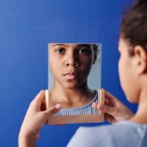 A young girl holds a mirror, reflecting her face, set against a vivid blue background.